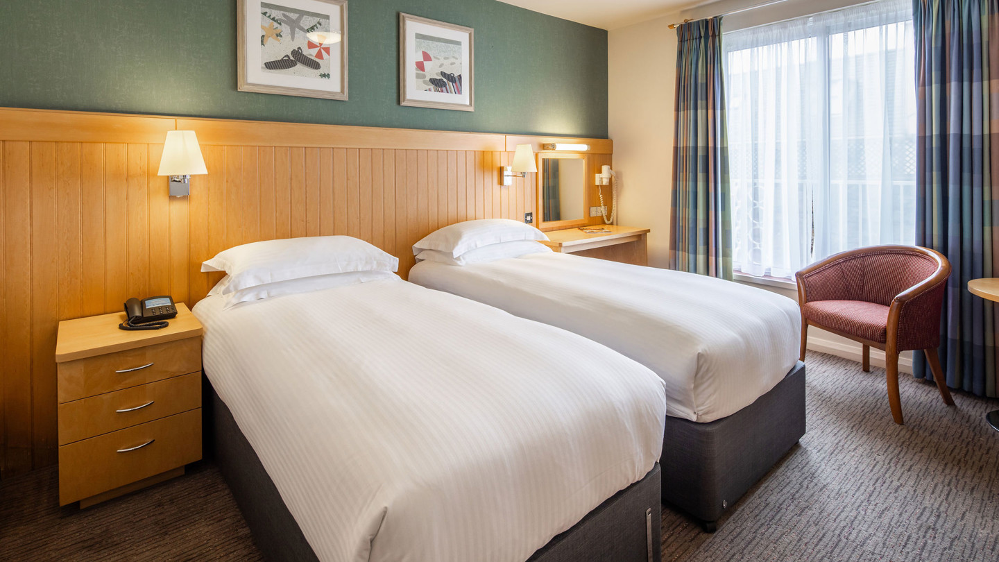 Two twin beds in a hotel room with crisp white linen, bed side tables and a small round table with armchairs